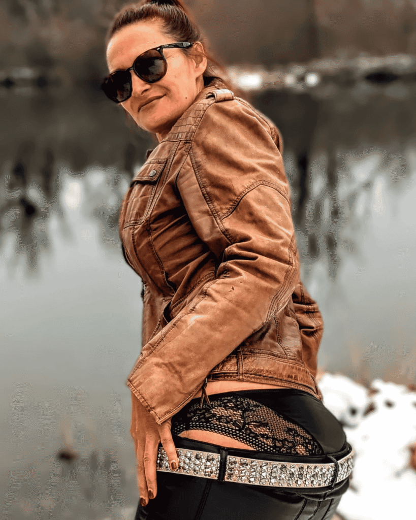 Letty Bixx steht in schwarzem Lederlook und brauner Lederjacke selbstbewusst am Wasser und verbindet rebellische Outdoor-Vibes mit moderner femininer Sinnlichkeit.